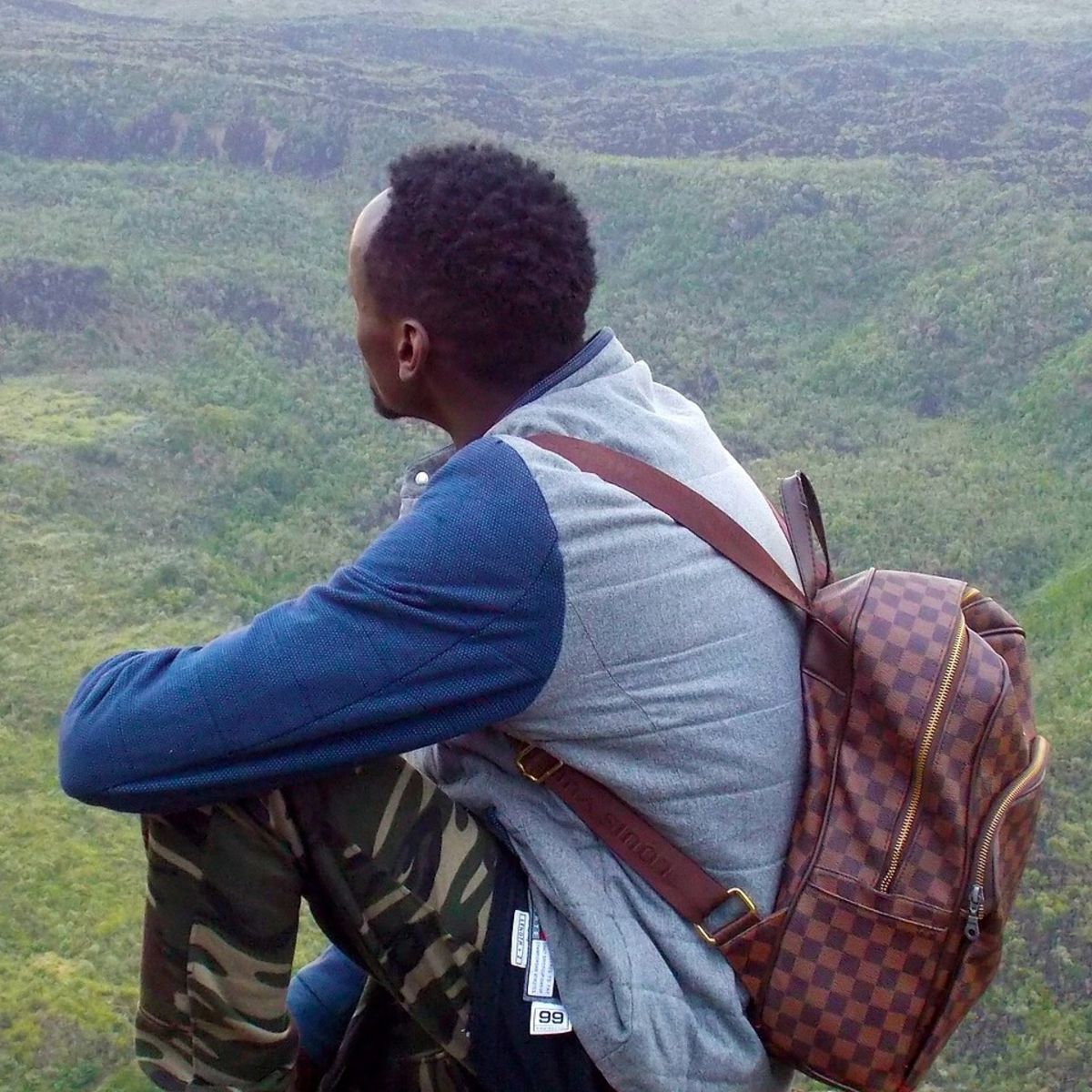 Young African man in yellow boots and wearing a checked backpack sitting on rock high above green landscape