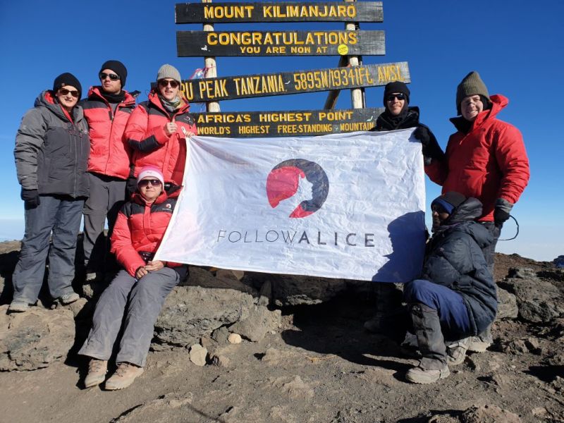 Group photo with Follow Alice flag at the summit sign for Uhuru Peak on Kilimanjaro