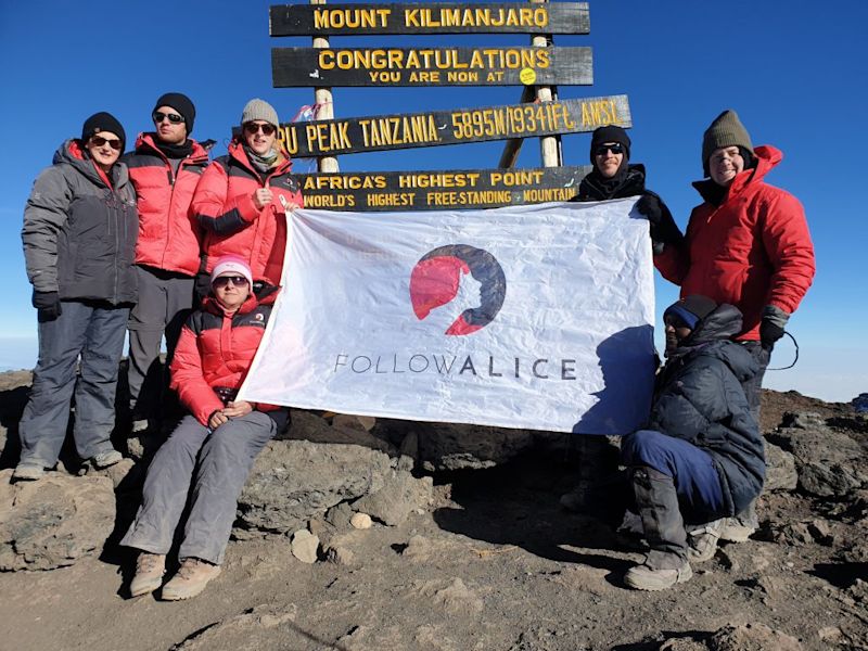 Group photo with Follow Alice flag at the summit sign for Uhuru Peak on Kilimanjaro