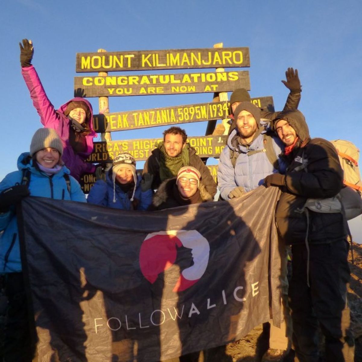 Group photo at Uhuru Peak on a sunny day with the Follow Alice flag
