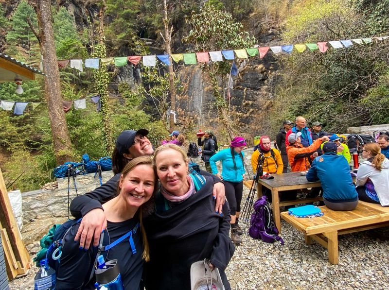Josh and Heather, April 2023. Two smiling female friend on EBC trek, early on, with trekkers sitting at outside table for rest break, Nepal