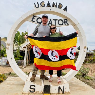 Couple smiling and holding Ugandan flag by Equator sign