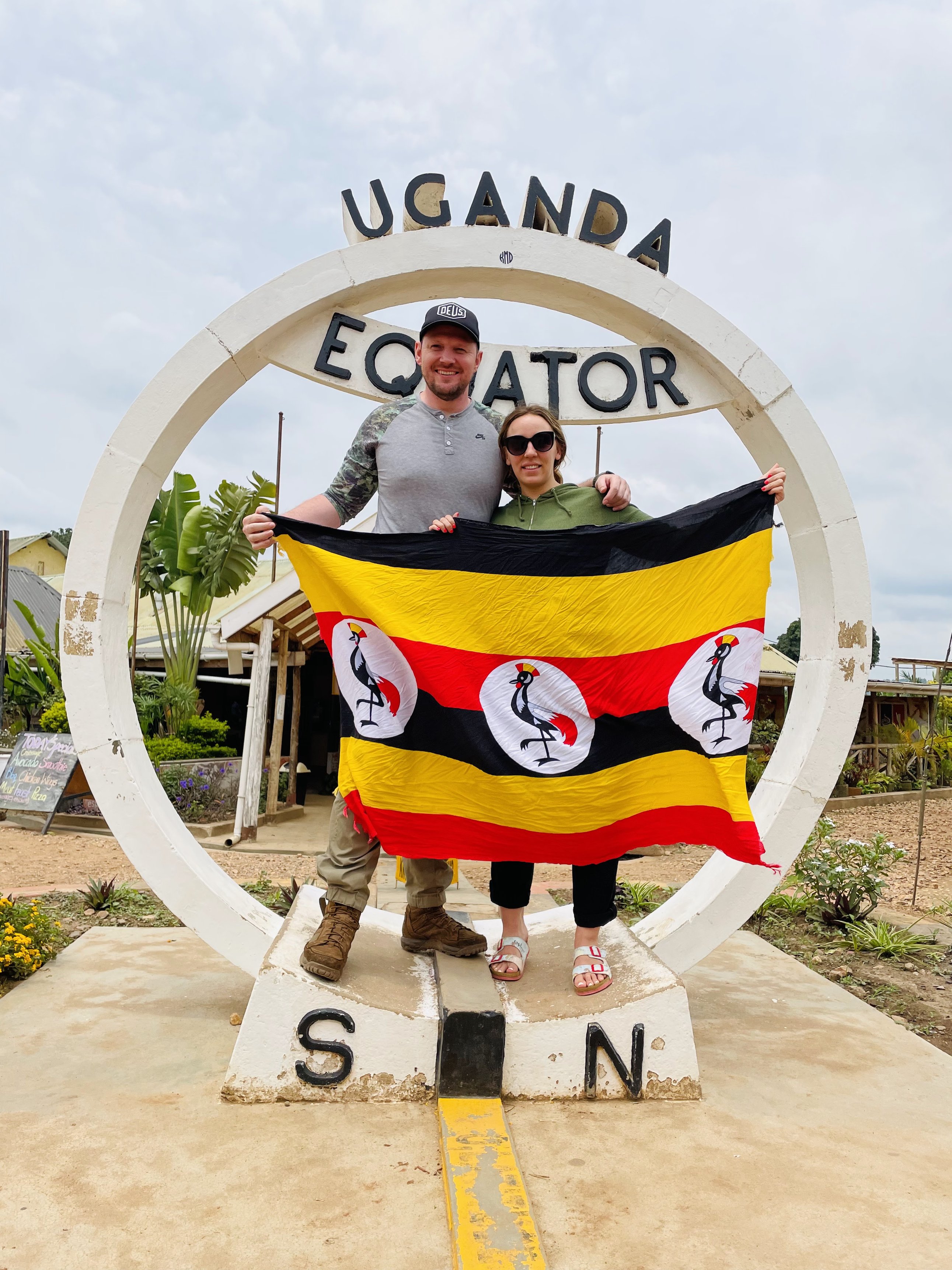 Couple smiling and holding Ugandan flag by Equator sign