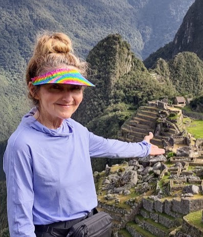 Woman trekker in hat and blue top pointing to Machu Picchu ruins behind her