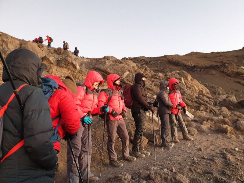 A line of Kilimanjaro trekkers in red Follow Alice jackets looking out over the view with the sunset light on them