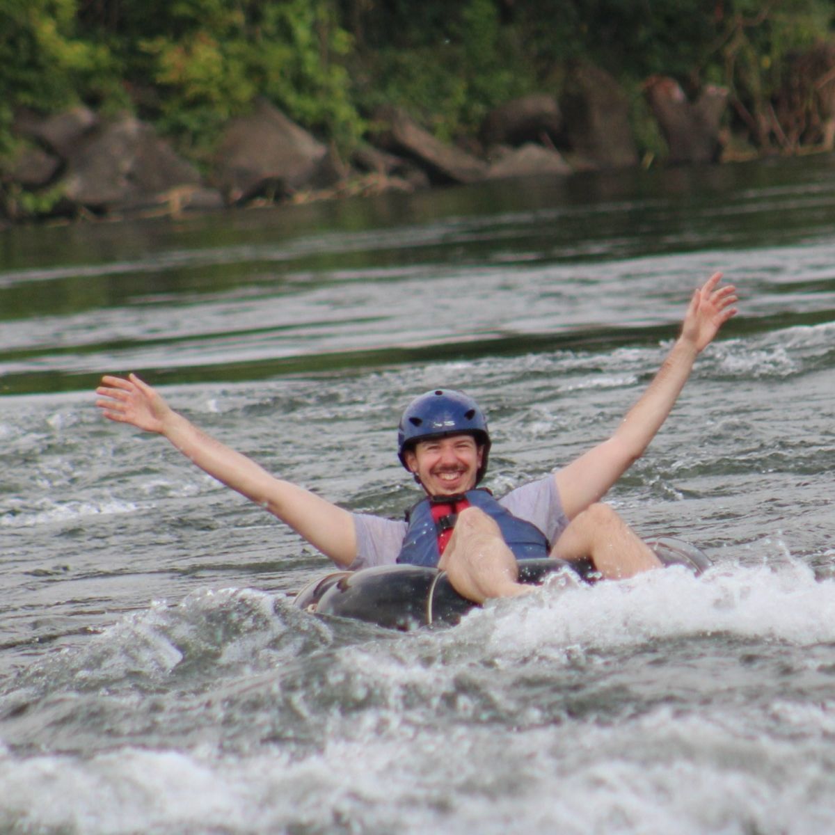 Man in hard hat with arms raised as he moves down river on the Nile while sitting in an inflated tube